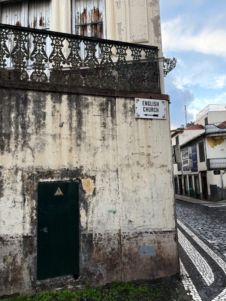 Łuszcząca się ściana starej kamienicy z kutym balkonem i zielonymi drzwiami, na rogu biała tabliczka English Church, obok brukowana uliczka