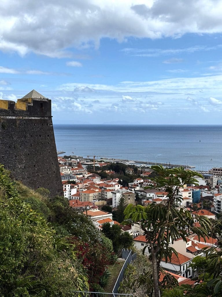 Narożna strażnica Fortaleza do Pico z piramidalnym daszkiem nad zielonym zboczem, poniżej dachy centrum Funchal i falochron portu na tle oceanu