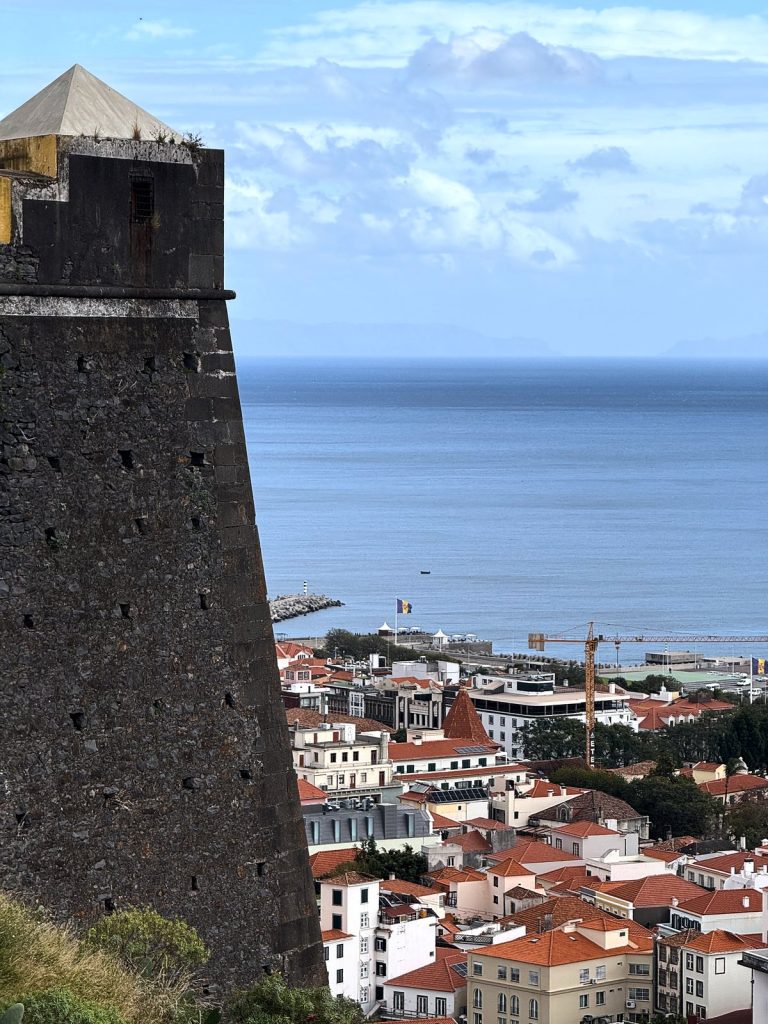 Ciemna kamienna baszta fortu z piramidalnym daszkiem, obok widok z góry na czerwone dachy Funchal, port i ocean