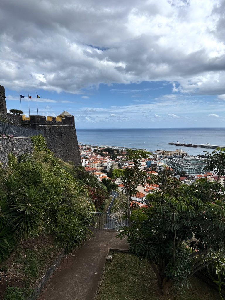 Ścieżka wśród zieleni pod murami Fortaleza do Pico, na murach trzy flagi i narożna strażnica z żółtymi blankami, w tle dachy Funchal, port i ocean