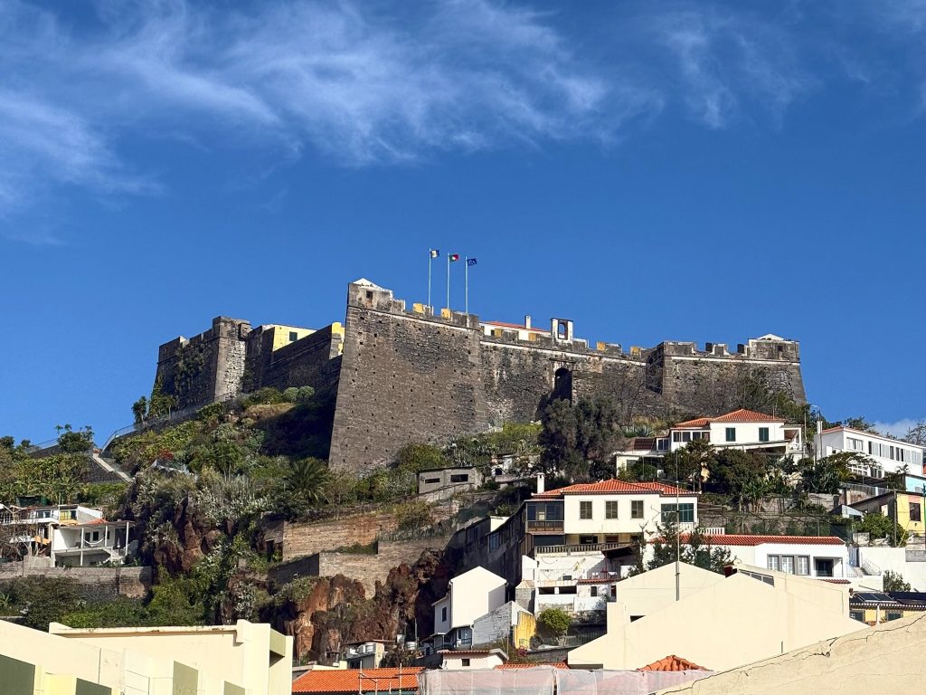 Fortaleza do Pico – ciemne bazaltowe mury twierdzy z trzema flagami na szczycie wzgórza, poniżej domy z czerwonymi dachami, nad nimi błękitne niebo z pierzastymi chmurami