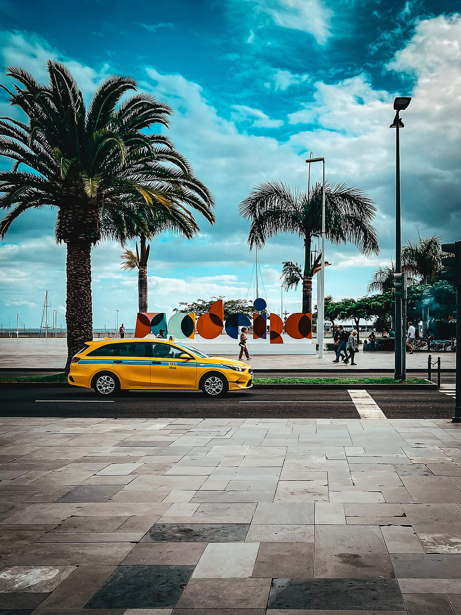 Żółta taksówka na Avenida do Mar w Funchal, w tle palmy i kolorowy napis Madeira