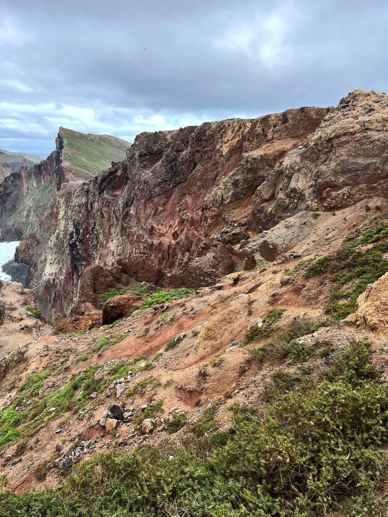 Strome czerwono-brunatne zbocze klifu z zielonymi kępami roślin, w tle skalisty grzbiet nad oceanem