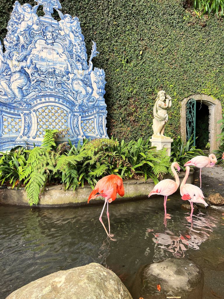 Różowe flamingi w stawie przed niebiesko-białym panelem azulejos i białym posągiem w ogrodzie Monte Palace