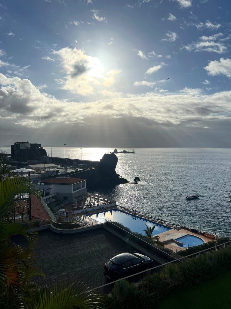 Słońce przebija się przez chmury nad oceanem, w dole hotelowe baseny, falochron i skała w morzu w zachodniej części Funchal