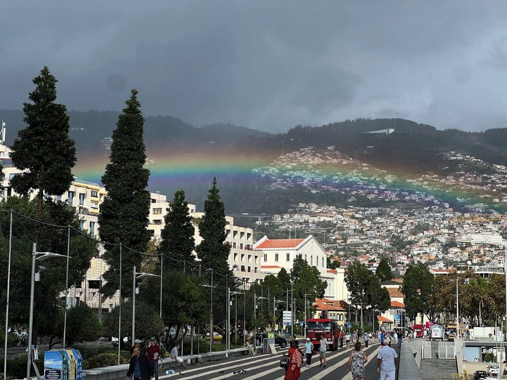 Tęcza nad zboczami Funchal pod ciemnymi chmurami, w dole promenada z drzewami, budynkami i drobnymi sylwetkami przechodniów