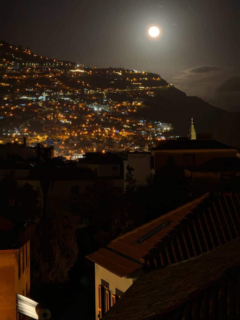Księżyc w pełni nad zboczem Funchal pełnym świateł, na pierwszym planie ciemne dachy