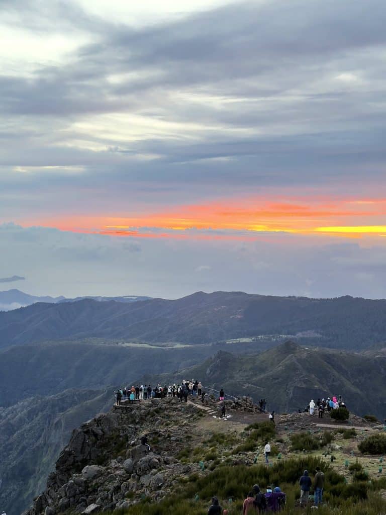Ludzie na skalistym punkcie widokowym na Pico do Arieiro o świcie, nad grzbietami gór pomarańczowy pas nieba