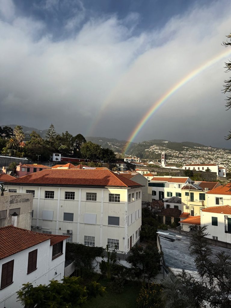 Podwójna tęcza nad dachami Funchal pod ciemnymi chmurami deszczowymi, na pierwszym planie biały budynek z pomarańczowym dachem, w tle zabudowa na zboczach