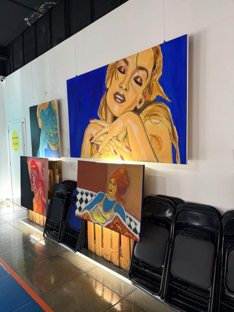 Duże kolorowe obrazy w stylu pop-art na białej ścianie korytarza, pod nimi oparte płótna i złożone krzesła