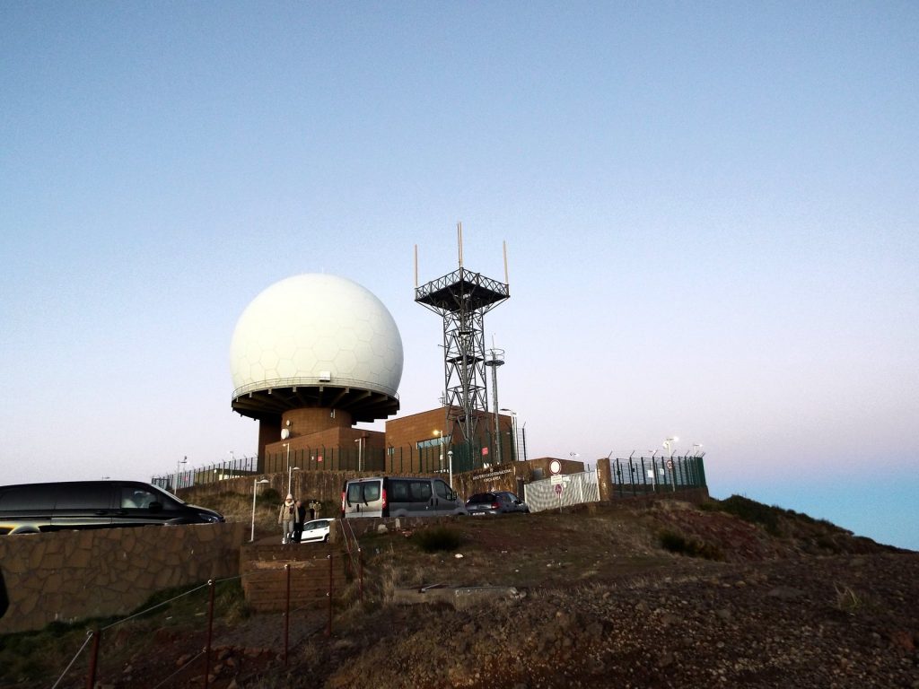 Kopuła radaru i stalowy maszt z antenami na szczycie Pico do Arieiro, przy budynkach zaparkowane samochody, różowe niebo o świcie