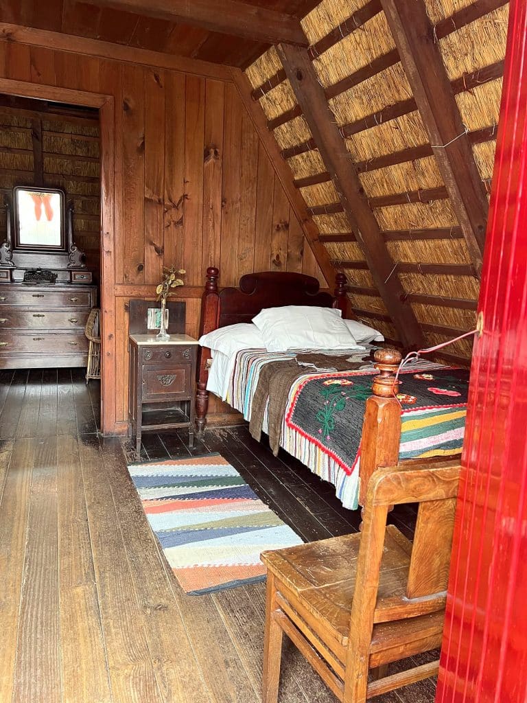 Wnętrze domku pod strzechą: drewniane łóżko z kolorową narzutą, chodnik w paski, krzesło i komoda z lustrem
