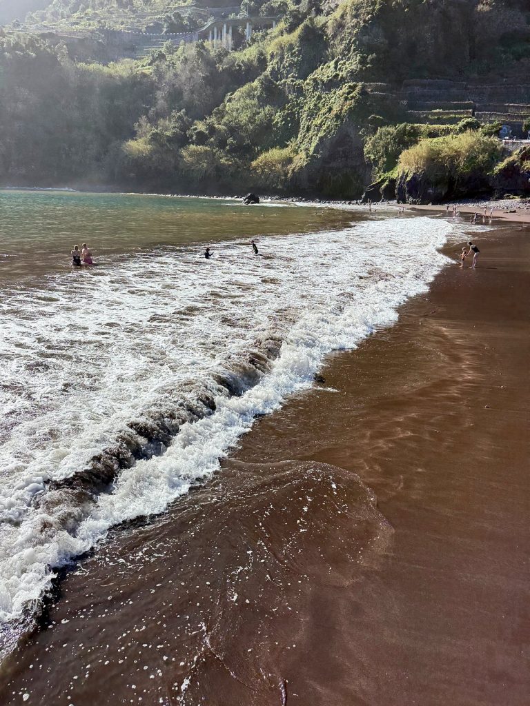 Spieniona fala wbiega na ciemny piasek plaży, w tle zielone klify w lekkiej mgiełce i kilka osób w wodzie