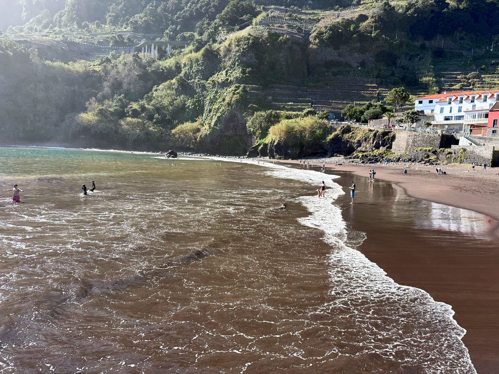 Fale wpływające na plażę z ciemnego piasku, w tle zielone zbocza i białe domy, w wodzie drobne sylwetki kąpiących się