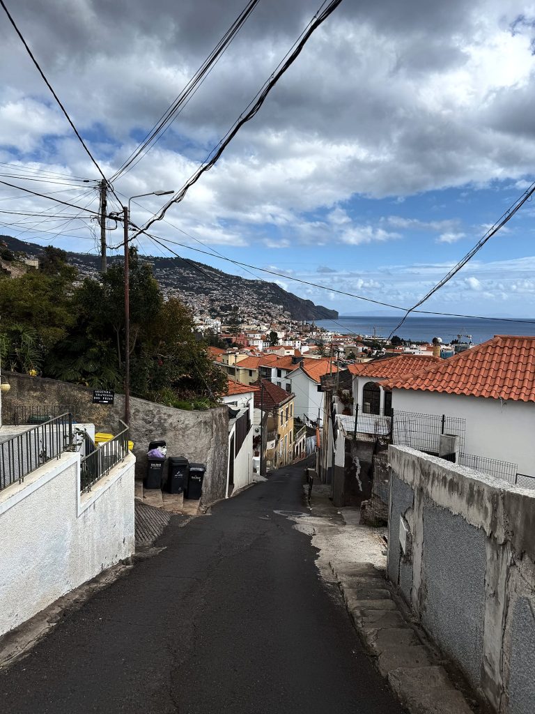 Stroma, wąska uliczka między białymi murami nad Funchal, w dole domy z pomarańczowymi dachami, ocean i zielone klify zachodniego wybrzeża