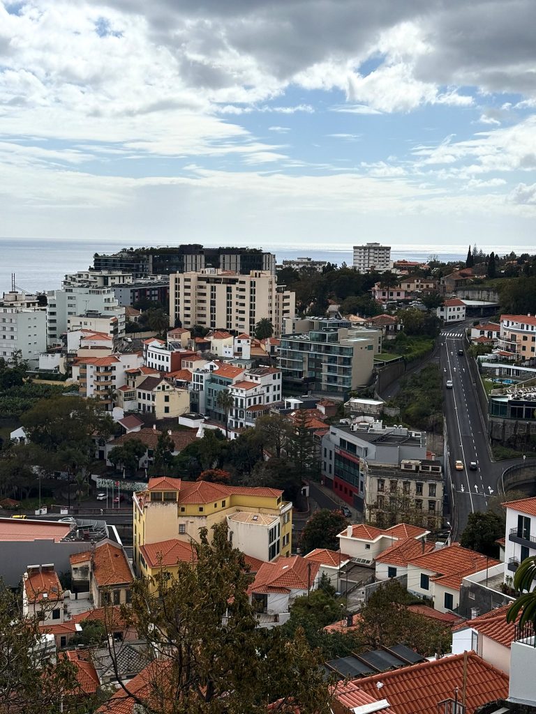 Widok z góry na zachodnią część Funchal: kamienice z pomarańczowymi dachami, ulica schodząca w dół, wysokie hotele i ocean pod zachmurzonym niebem